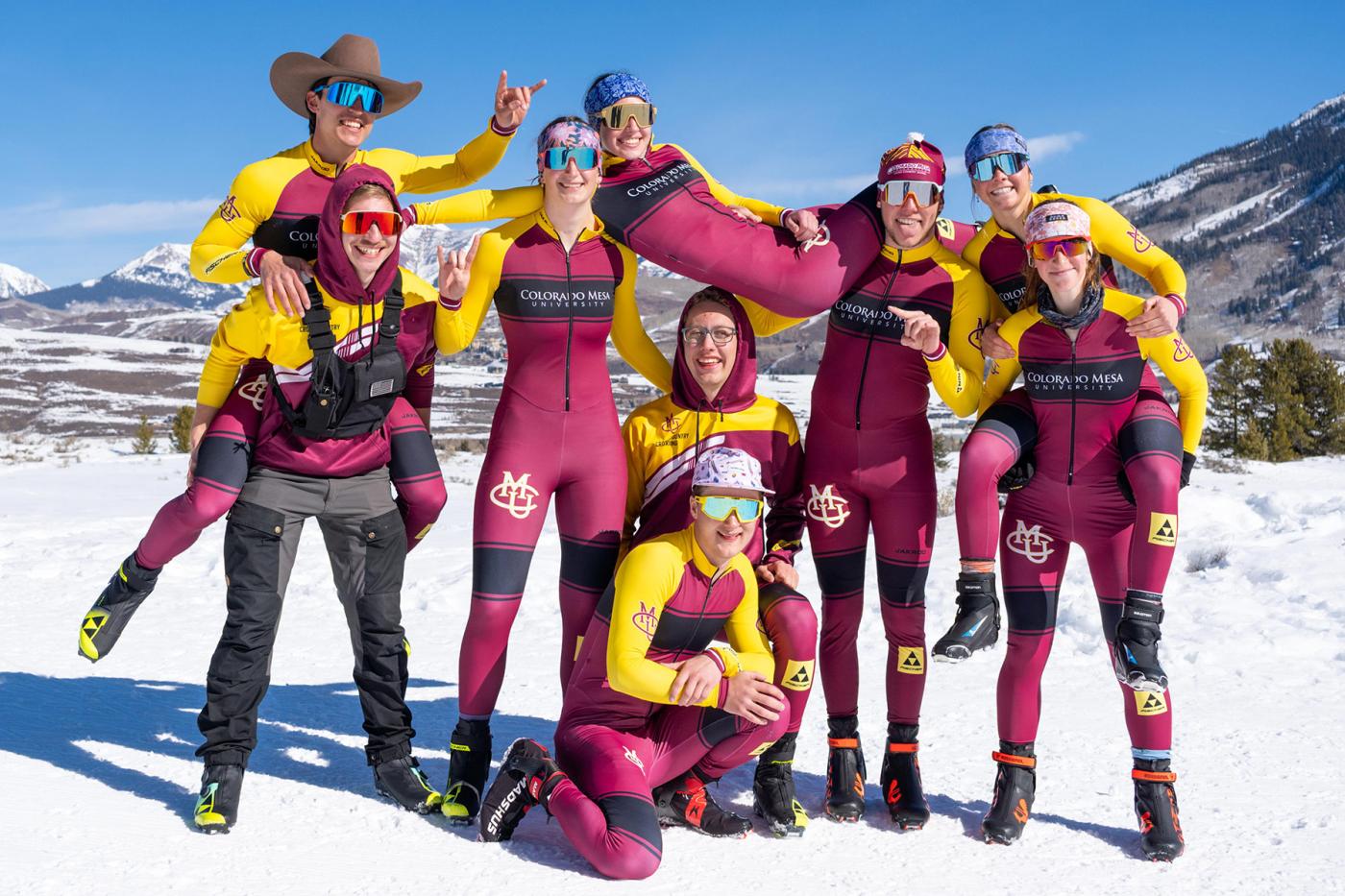 CMU Cross Country Ski Team posing together in matching maroon and gold race suits on a snowy mountain landscape, smiling and celebrating as a close-knit group.