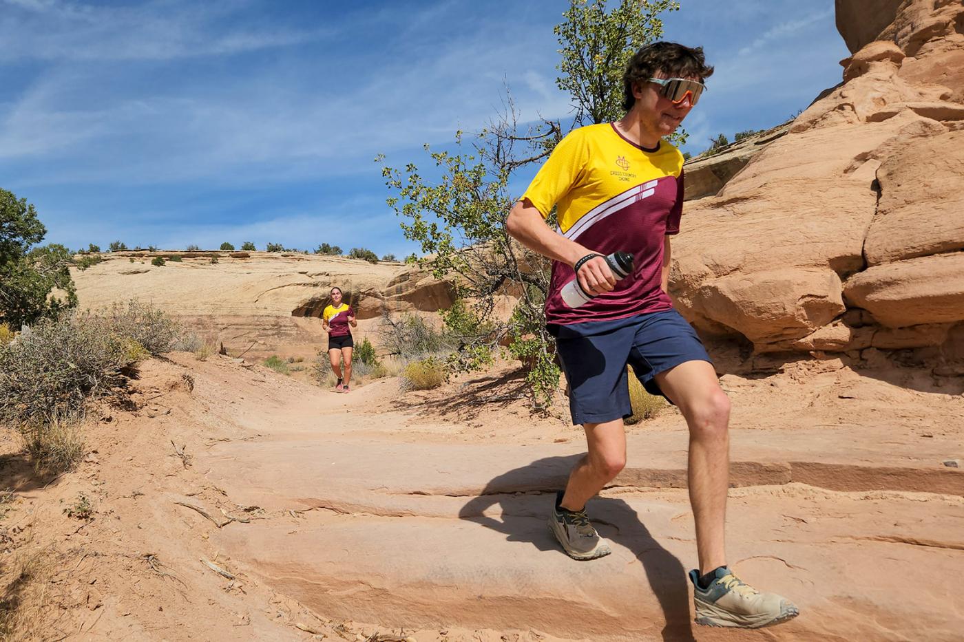 CMU Nordic ski team athletes trail running through a sandstone desert canyon near Grand Junction, Colorado during summer endurance training. One athlete descends a rocky section of trail while another follows behind under a blue sky.